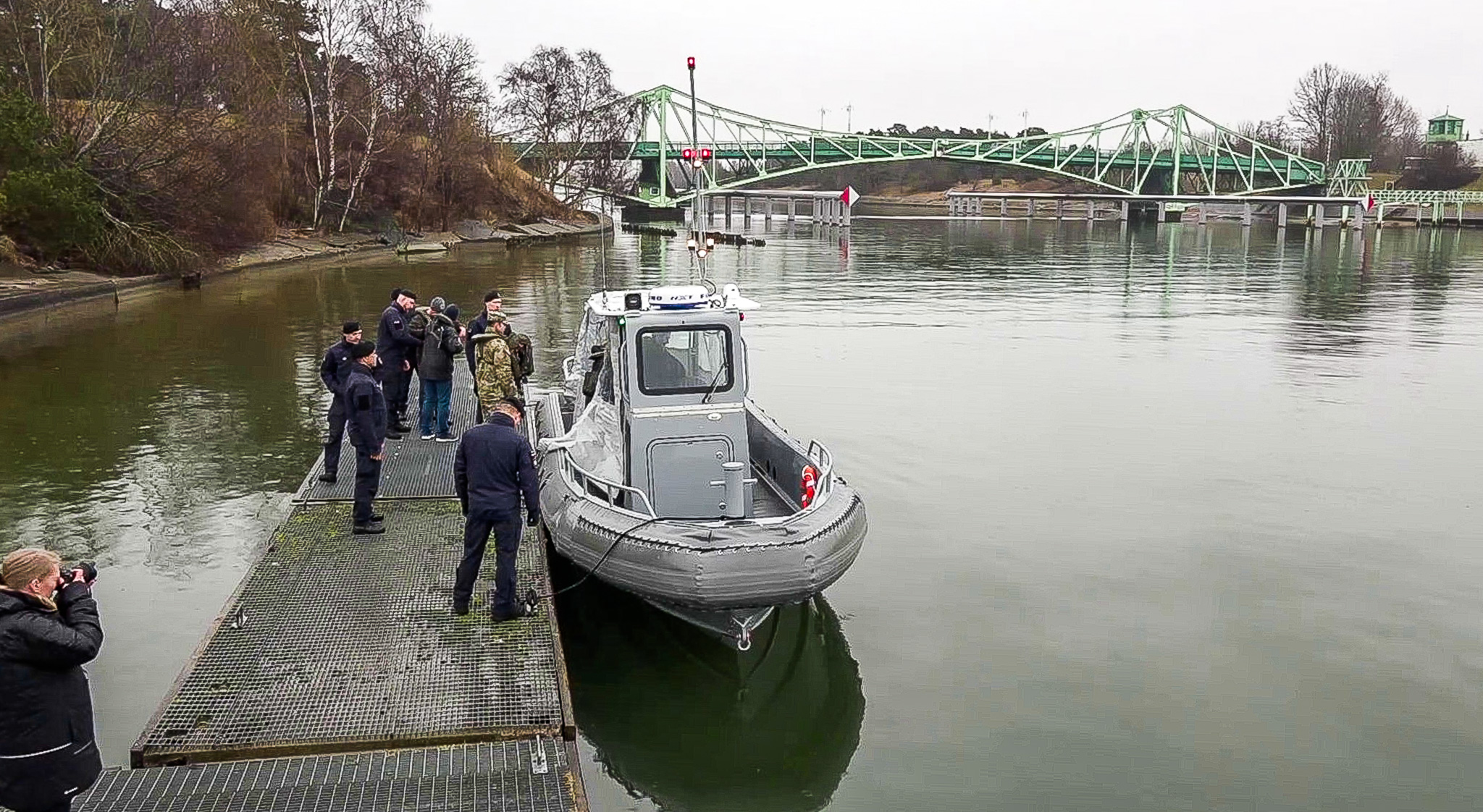 Pelēkas krāsas kuteris pie piestātnes ar uniformētiem virsniekiem, fonā redzams zaļš tilts un krasta ainava