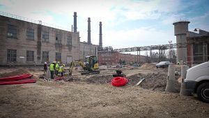 Būvniecības darbi industriālā teritorijā ar ekskavatoru un strādniekiem pie vecām rūpnīcu ēkām