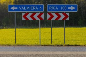 Ceļa zīmes ar norādēm “Valmiera 6 km” un “Rīga 100 km” uz asfalta ceļa ar zaļu pļavu fonā – kadrs no skrējiensoļojuma Rīga–Valmiera.