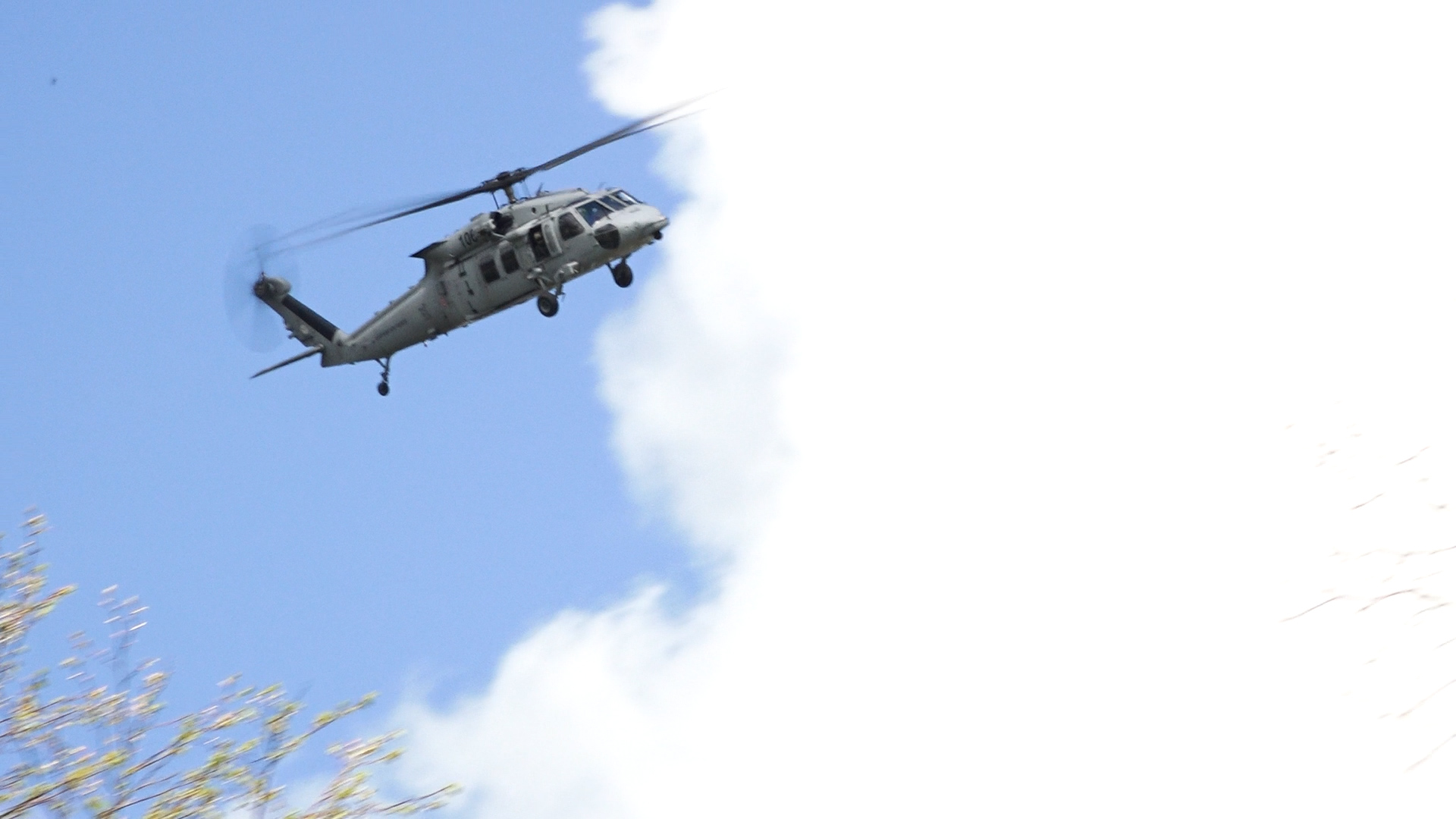Militārais helikopters lido virs apvidus ar ziliem debesīm un mākoņiem fonā