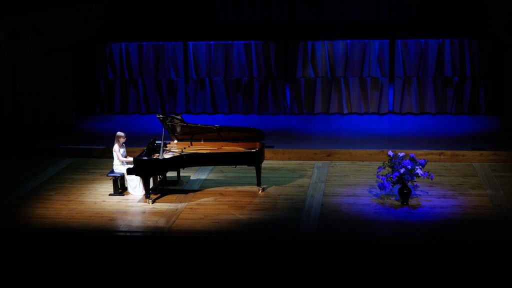 Pianiste uz plašas skatuves ar flīģeli un ziediem