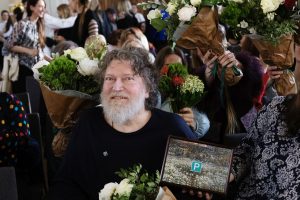 Mākslinieks Romāns Korovins ar bārdu un sirmiem matiem smaidot sēž ar balvu un ziedu pušķiem apkārt