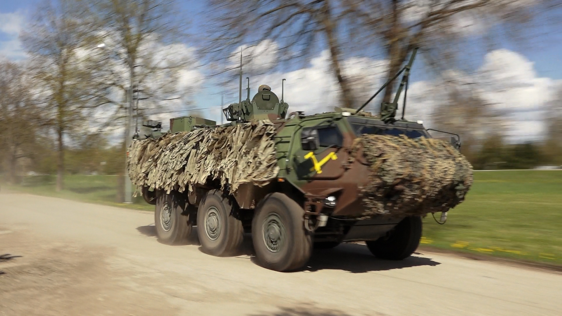 Maskēts militārais transportlīdzeklis kustībā pa grants ceļu apvidū ar kokiem