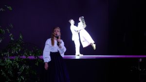 Dziedātāja uzstājas uz skatuves, fonā dejo pāris romantiskā apgaismojumā