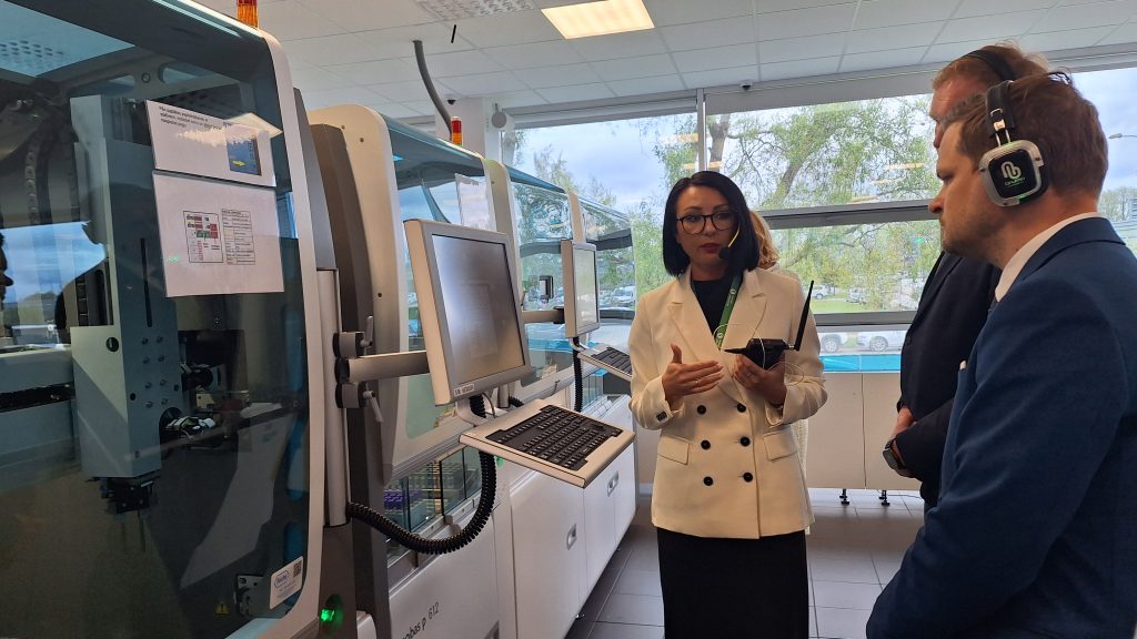 Sieviete baltā žaketē stāsta diviem vīriešiem par automatizēto laboratorijas iekārtu