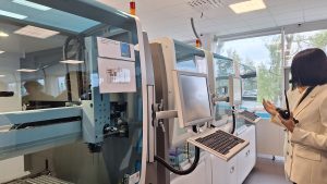 Sieviete baltā žaketē rāda uz automatizēto laboratorijas iekārtu