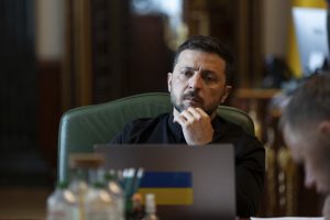 Ukrainas prezidents Volodimirs Zelenskis ar domīgu sejas izteiksmi sēž pie datora ar Ukrainas karoga uzlīmi