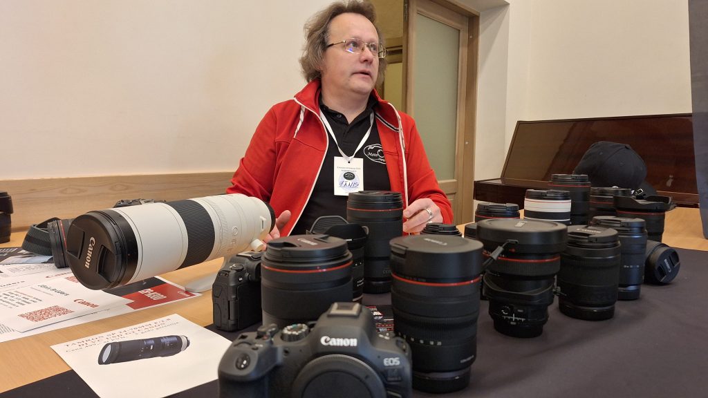 Vīrietis ar brillēm sēž pie galda, ko klāj dažādas fotokameras un objektīvi