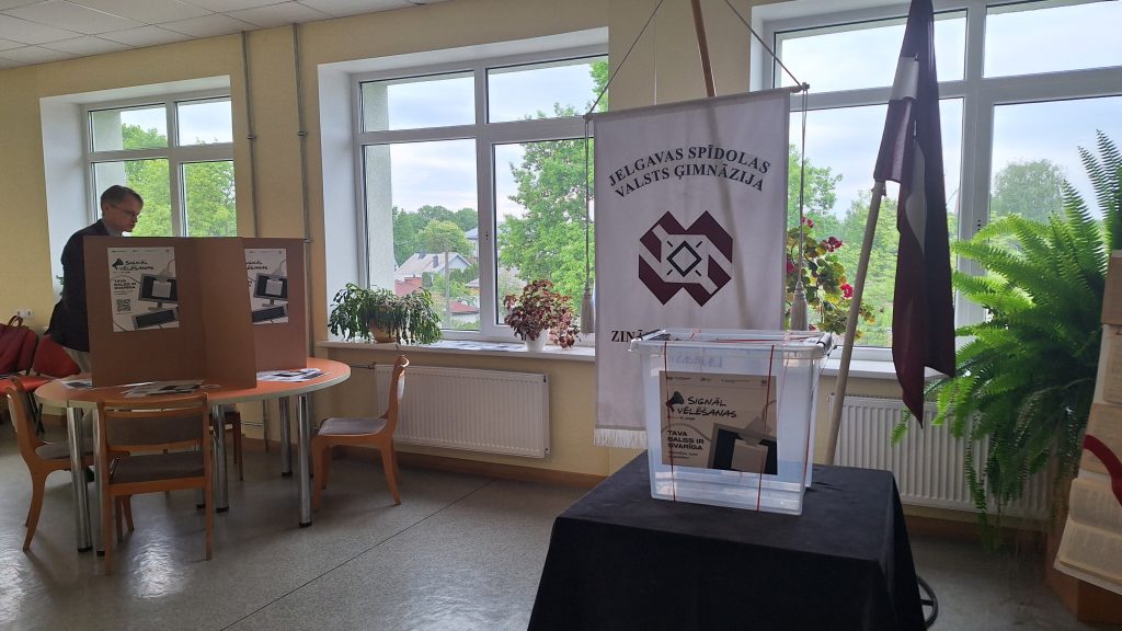 Vēlēšanu urna un Jelgavas Spīdolas Valsts ģimnāzijas karogs vēlēšanu iecirknī