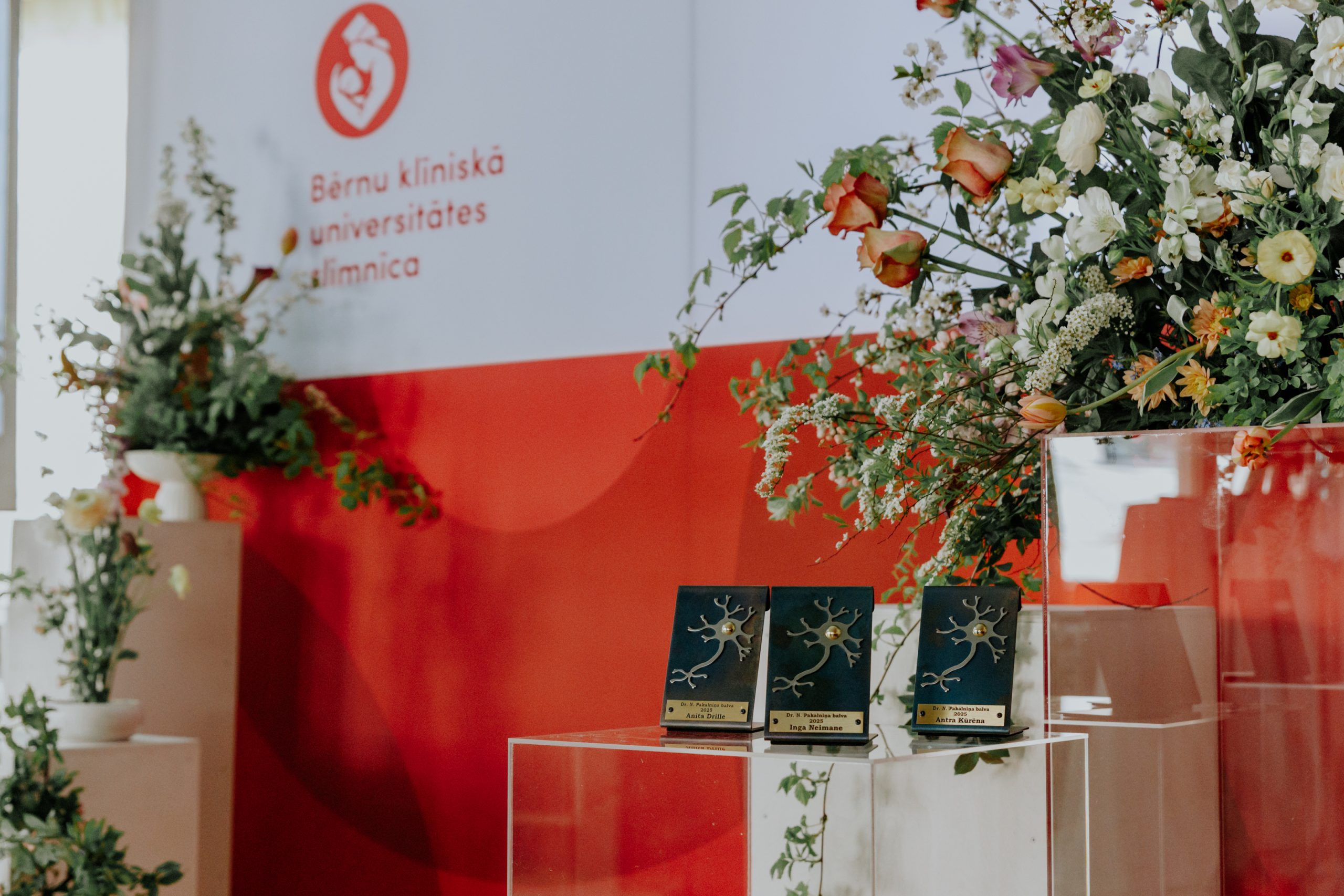 Nikolaja Pakalniņa balvas uz stikla pjedestāla ar Bērnu slimnīcas logotipu un ziedu dekorācijām fonā.