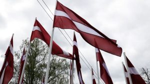 Latvijas karogi plīvo debesīs pret pelēku mākoņu fonu
