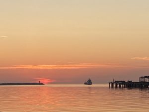 Saulriets Ventspils ostā ar kuģi pie apvāršņa un piestātni priekšplānā