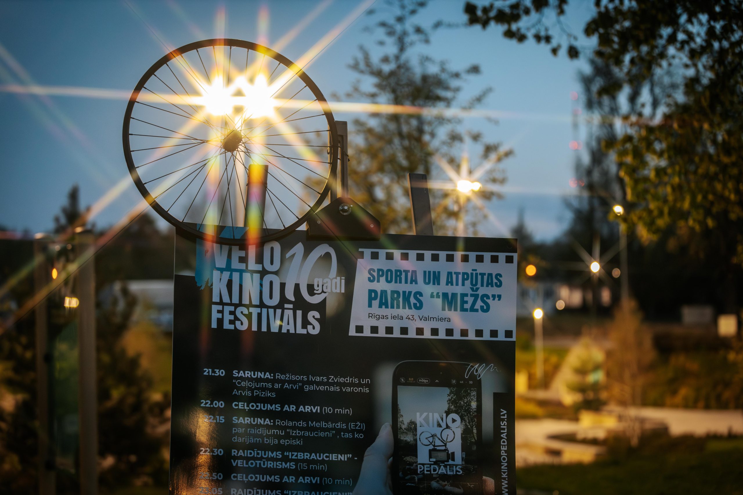 Velo kino festivāla Kinopedālis plakāts Valmierā ar riteni un starojošu apgaismojumu fonā.