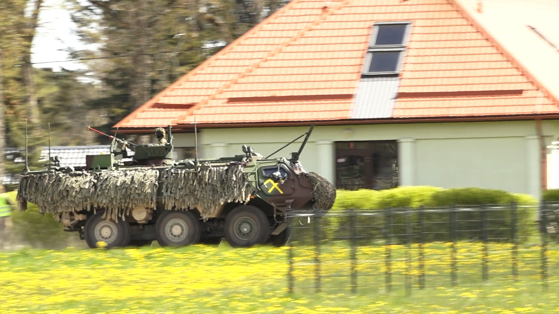 Maskēts militārais transportlīdzeklis pārvietojas pa ziedošu pļavu apdzīvotā vietā