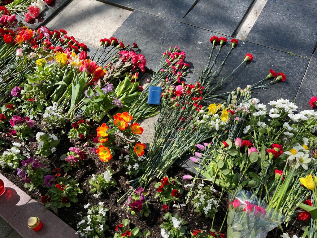 uvplānā redzami dažādi ziedi - sarkanas un dzeltenas tulpes, sarkanas neļķes, kā arī citi krāsaini ziedi, kas nolikti uz tumšām akmens plāksnēm un zemē. Redzamas arī dažas degošas svecītes sarkanos trauciņos.