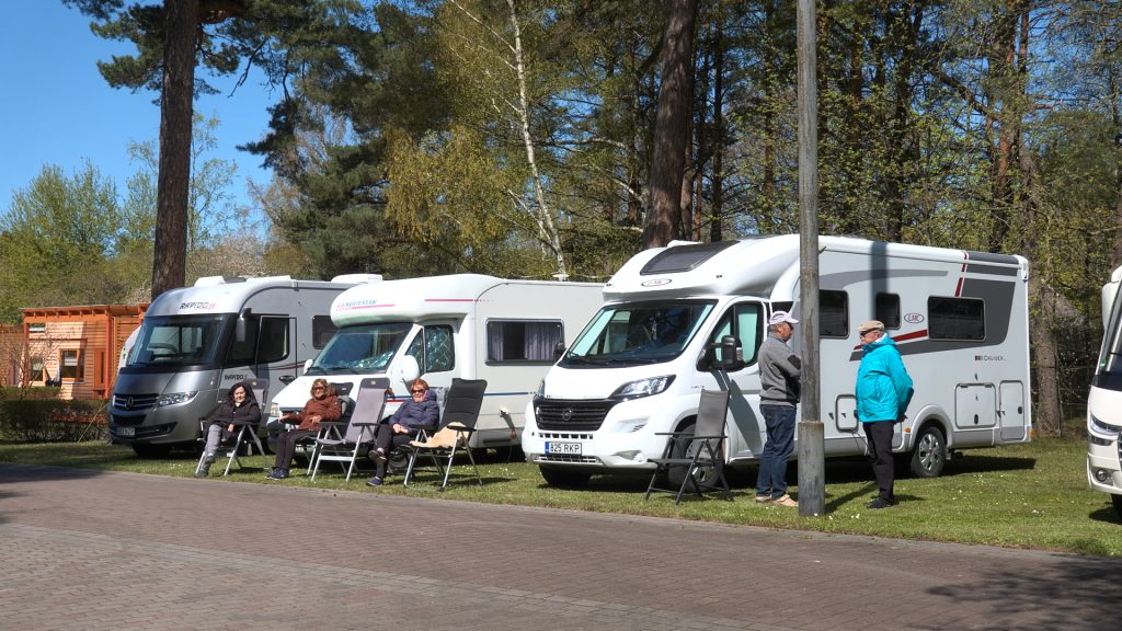 Vairāki kemperi novietoti stāvvietā Ventspils kempingā, cilvēki sēž saliekamajos krēslos.