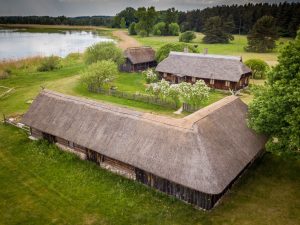 Tradicionāla Latvijas lauku sēta pie ezera ar niedru jumta mājām un ziedošiem kokiem