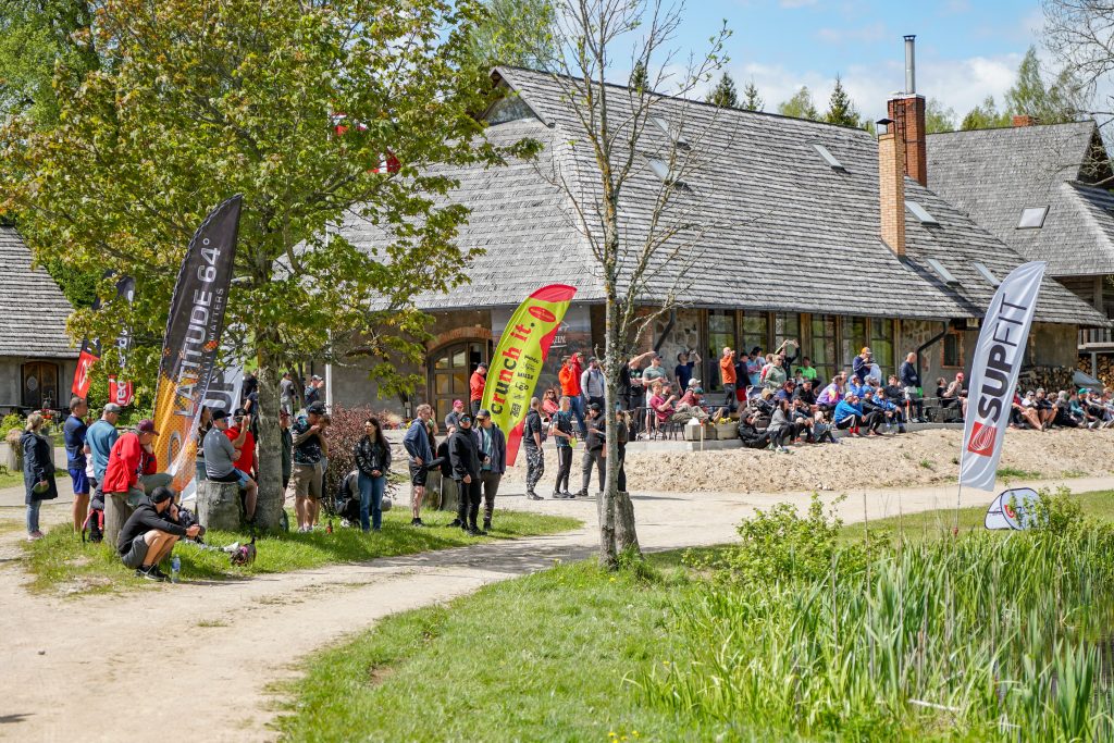 Dalībnieki pulcējas pie ēkas disku golfa festivāla norises vietā ar karogiem