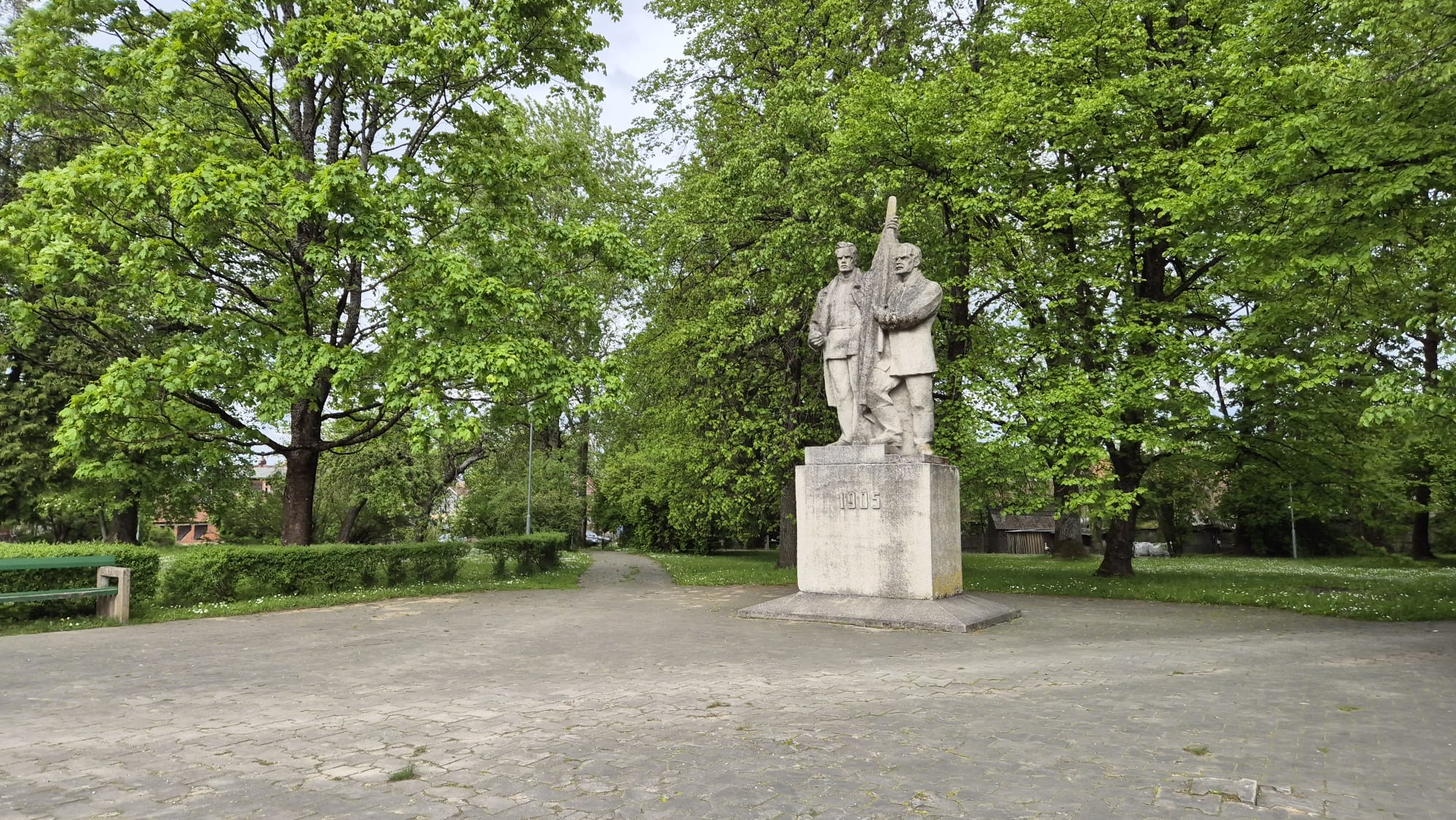 Piemineklis 1905. gada revolucionāriem Kuldīgas parkā, ko ieskauj koki un zaļumi, simbolizējot parka vēsturisko nozīmi