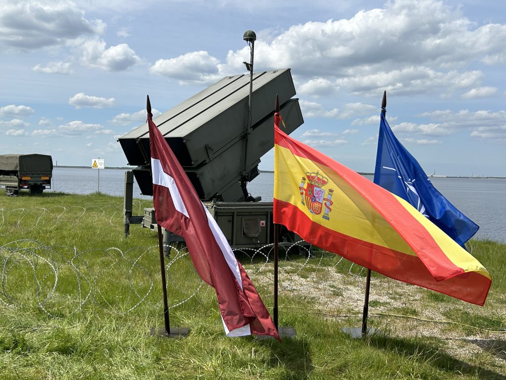 NATO pretgaisa aizsardzības sistēma NASAMS un militārais transportlīdzeklis ar Latvijas, Spānijas un NATO karogiem, novietoti pie ūdenstilpes mācību laikā