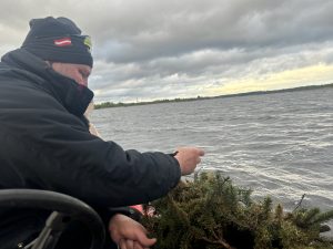Vīrietis cepurē ar Latvijas karoga emblēmu no laivas ievieto ūdenī mākslīgo nārstu ligzdu