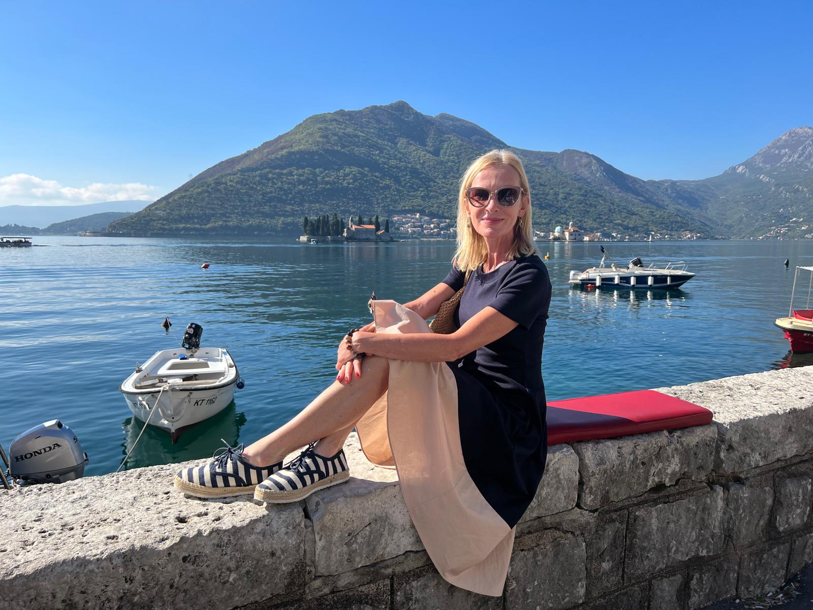 Ieva Keiša sēž pie jūras ar kalniem fonā, baudot saulainu dienu