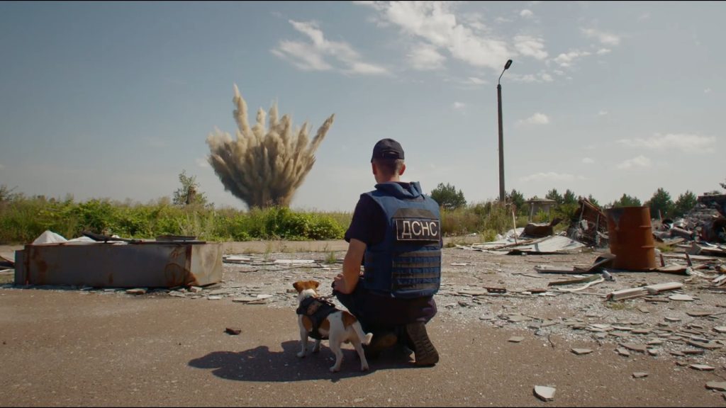 Vīrietis formas tērpā ar suni skatās uz tālu sprādzienu ainavā ar drupām
