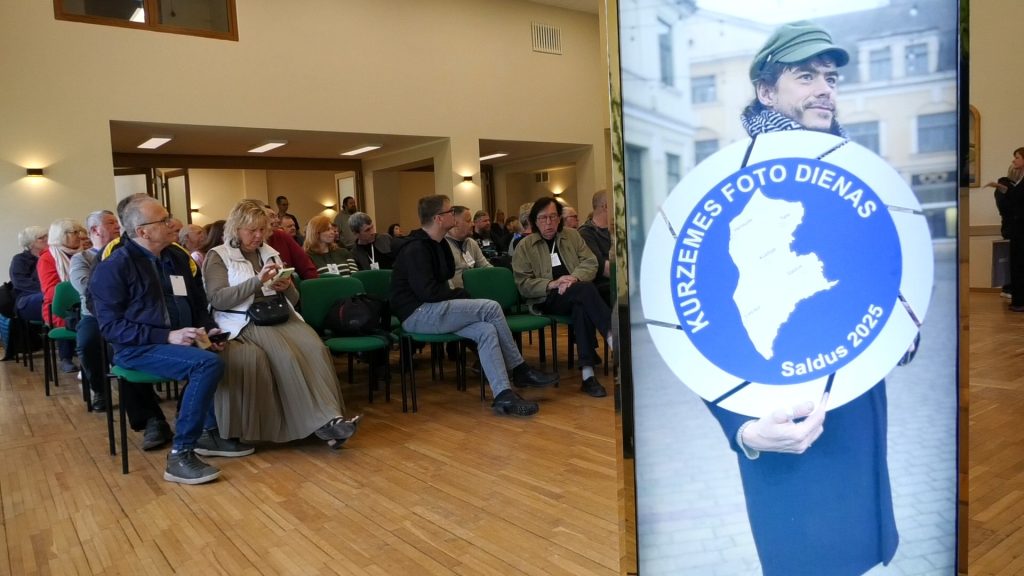 Cilvēki apmeklē Kurzemes foto dienas pasākumu, blakus stendam ar uzrakstu "Kurzemes foto dienas Saldus 2025"