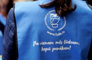 Persona zilā vestē ar LIZDA logo un saukli “Pa vienam mēs lūdzam, kopā panākam”