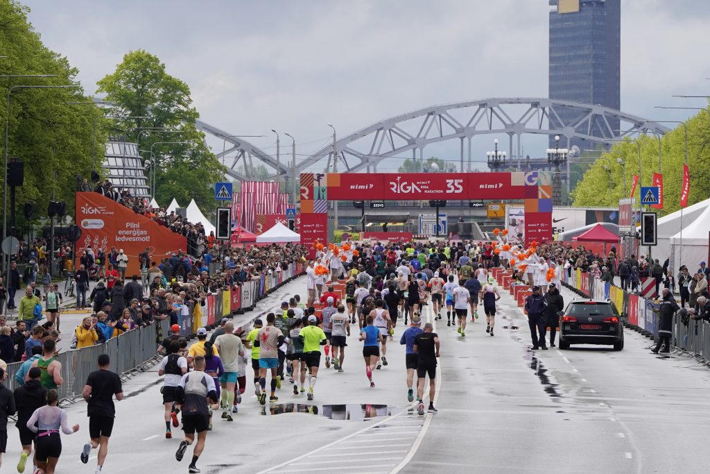 Dalībnieki skrien Rimi Rīgas maratona finišam tuvojošā posmā, fonā redzama Rīgas panorāma.