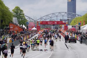 Dalībnieki skrien Rimi Rīgas maratona finišam tuvojošā posmā, fonā redzama Rīgas panorāma.