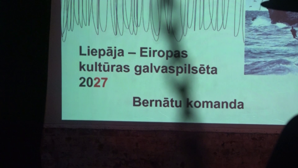 Projekcijas ekrāns ar uzrakstu "Liepāja - Eiropas kultūras galvaspilsēta 2027" un "Bernātu komanda".