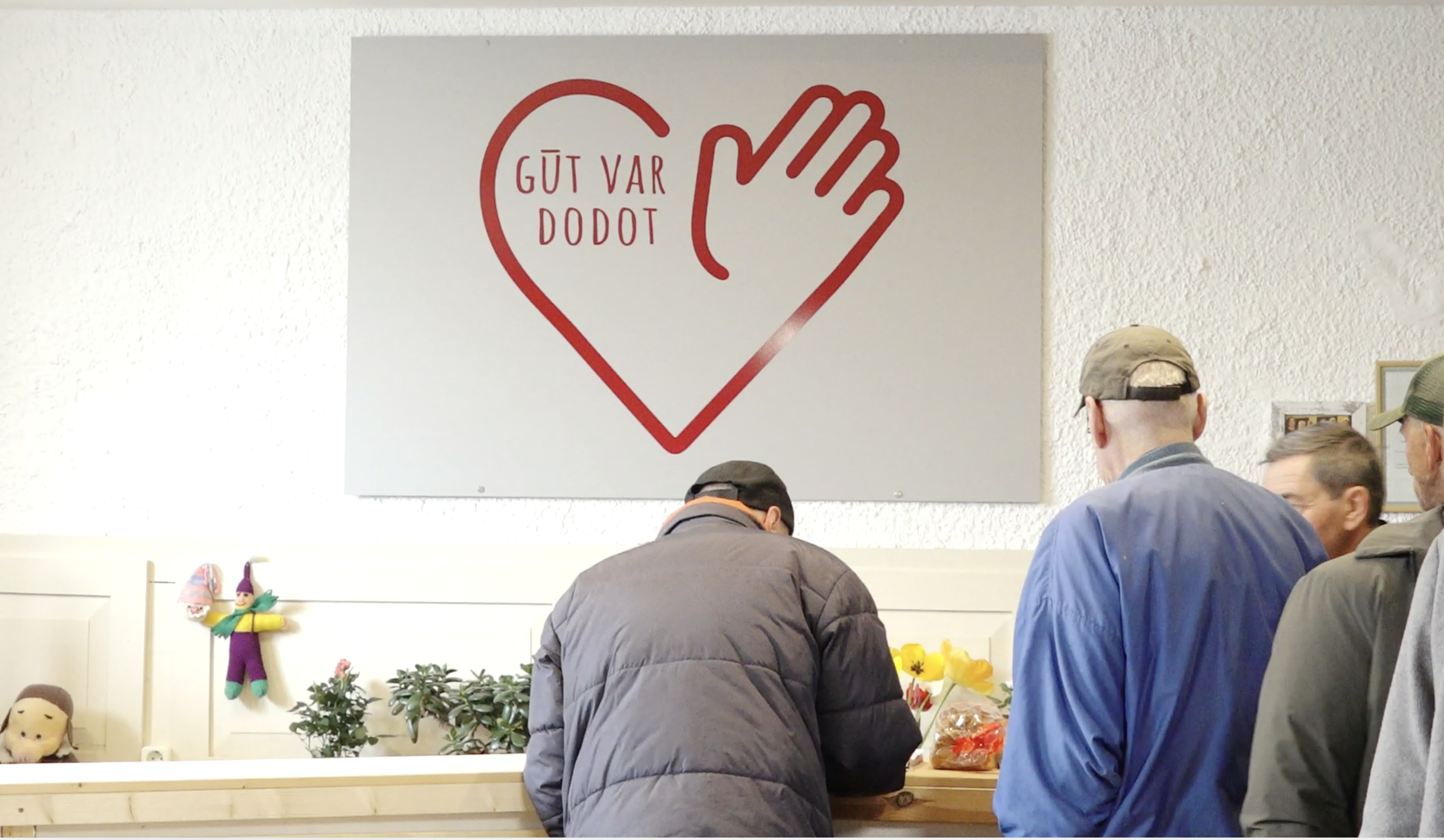 Cilvēki labdarības organizācijas telpā pie sienas ar uzrakstu “Gūt var dodot”