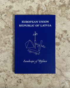 Latvijas paviljona brošūras vāks ar uzrakstu “Landscape of Defence” uz zilā fona.