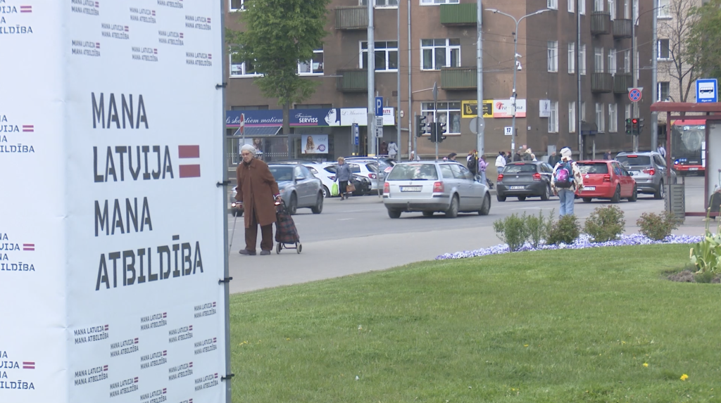 Gados vecāka sieviete ar iepirkumu ratiņiem staigā pa ielu Daugavpilī blakus plakātam "Mana Latvija = Mana Atbildība".