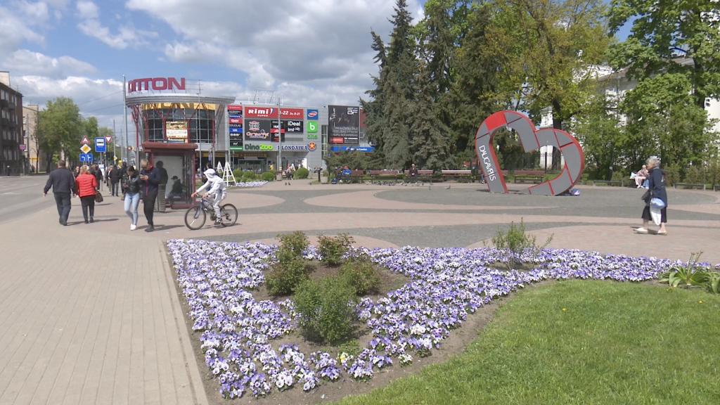 Cilvēki pastaigājas pa Daugavpils pilsētas laukumu ar tirdzniecības centru fonā un ziedu dobēm priekšplānā