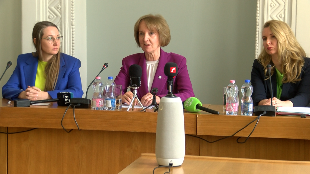 Trīs sievietes sēž pie galda preses konferencē, priekšplānā redzami mikrofoni, kas norāda uz publisku paziņojumu vai diskusiju