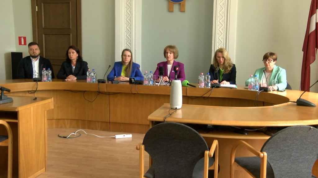Seši cilvēki sēž pie izliekta koka galda konferenču zālē, priekšplānā redzami mikrofoni