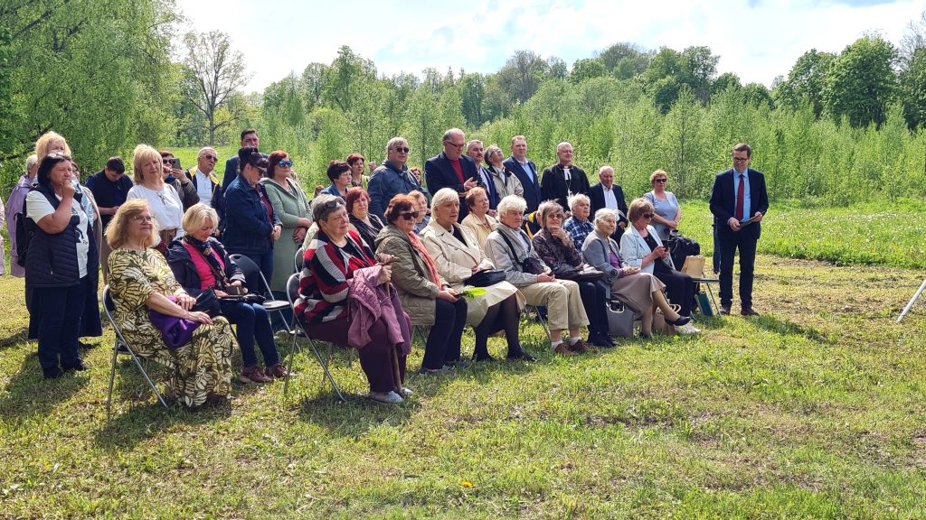 Cilvēku grupa sēž uz krēsliem un klausās uzrunu brīvdabas pasākumā