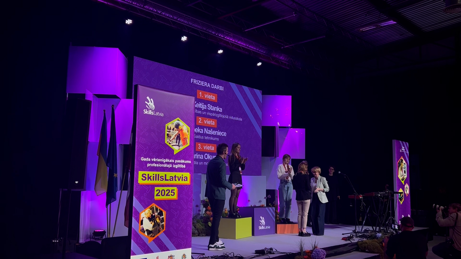 Trīs jaunietes stāv uz pjedestāla "SkillsLatvia 2025" frizieru darbu konkursa apbalvošanas ceremonijā