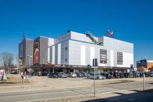 Tirdzniecības centrs “Valleta” Valmierā saulainā dienā, redzams fasādes skats un autostāvvieta priekšā.