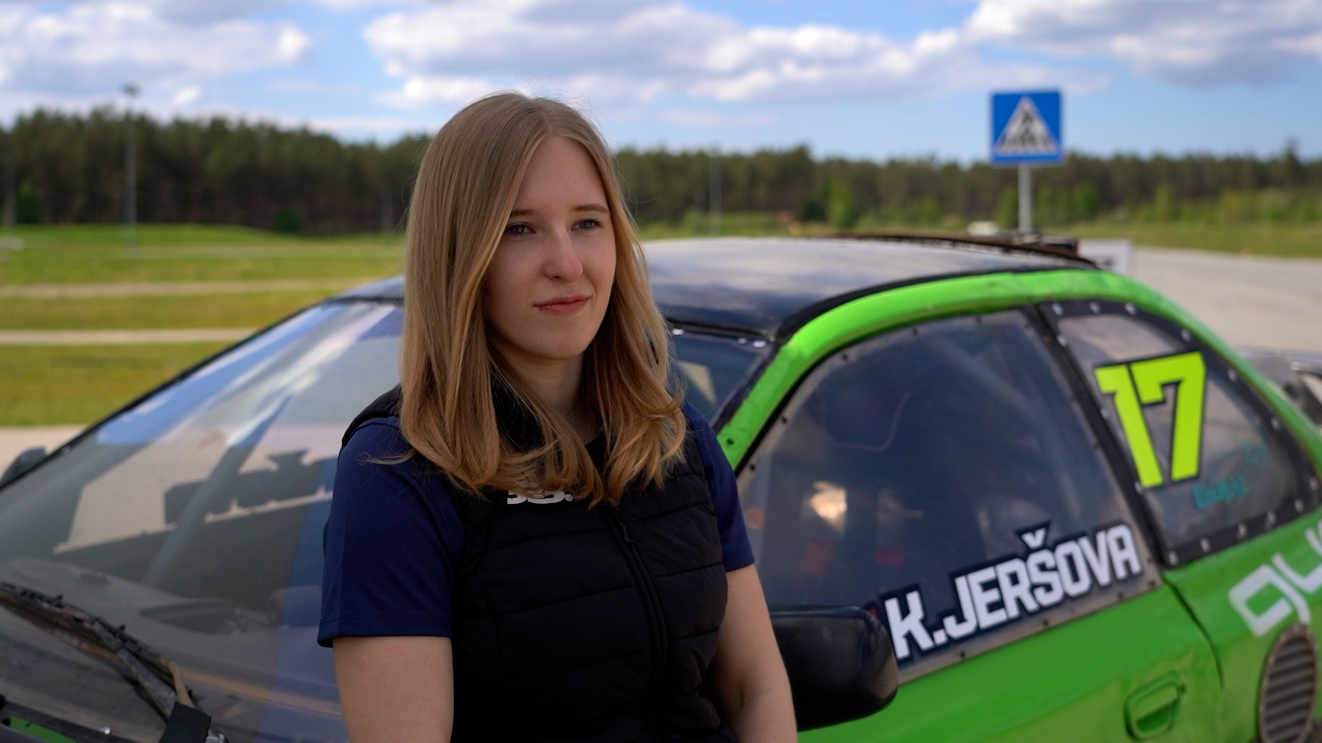 Autosportiste Kitija Jeršova stāv pie sava zaļā sacīkšu auto ar numuru 17