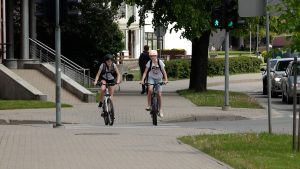 Divi bērni ar ķiverēm brauc ar velosipēdiem pa gājēju pāreju pie zaļās gaismas