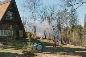 lauku māja ar niedru jumtu un cilvēki pie dūmiem pavasara darbos