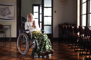 Sieviete ratiņkrēslā lūdzas klusā telpā ar koka grīdu un sakārtotiem krēsliem