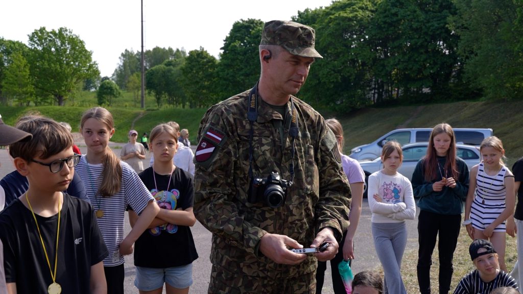 Vīrietis militārā formā stāv bērnu un jauniešu grupā stāsta par darbu Nacionālajos bruņotajos spēkos