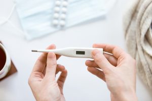 Rokas tur elektronisko termometru uz balta fona ar zālēm un tējas krūzi