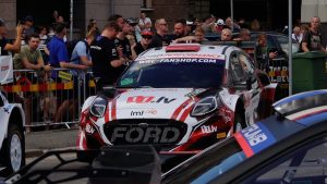 Ford rallija auto ar Latvijas reklāmām skatītāju un komandas locekļu ielokā