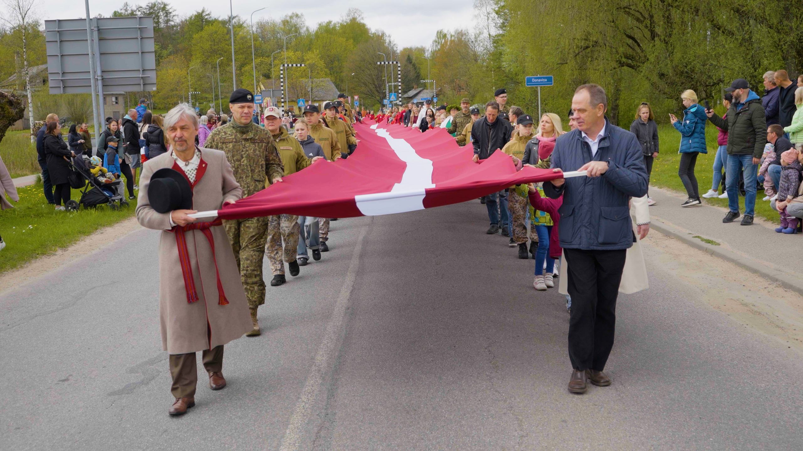 Cilvēki patriotiskā gājienā Limbažu ielās nes Latvijas valsts karogu, piedaloties karavīriem un civiliedzīvotājiem.