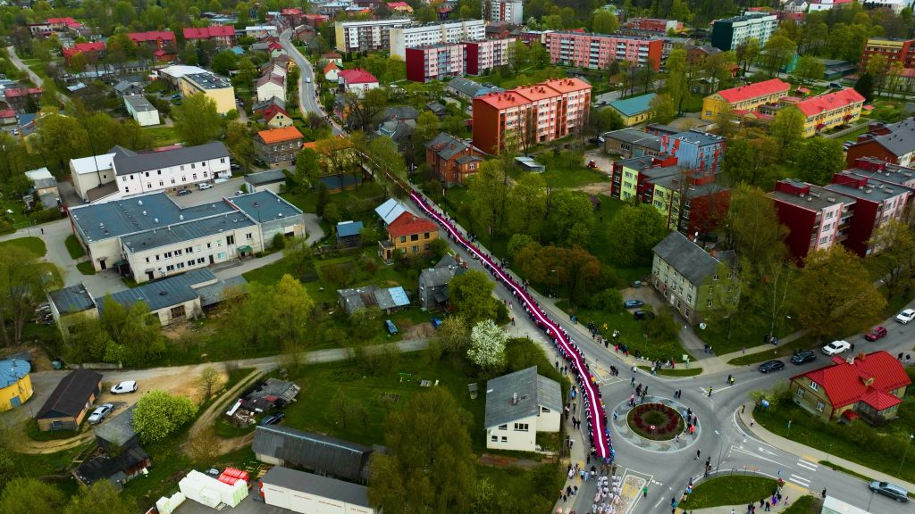 Gaisa skats uz cilvēku gājienu ar garu Latvijas valsts karogu, kas vijas cauri Limbažu ielām.
